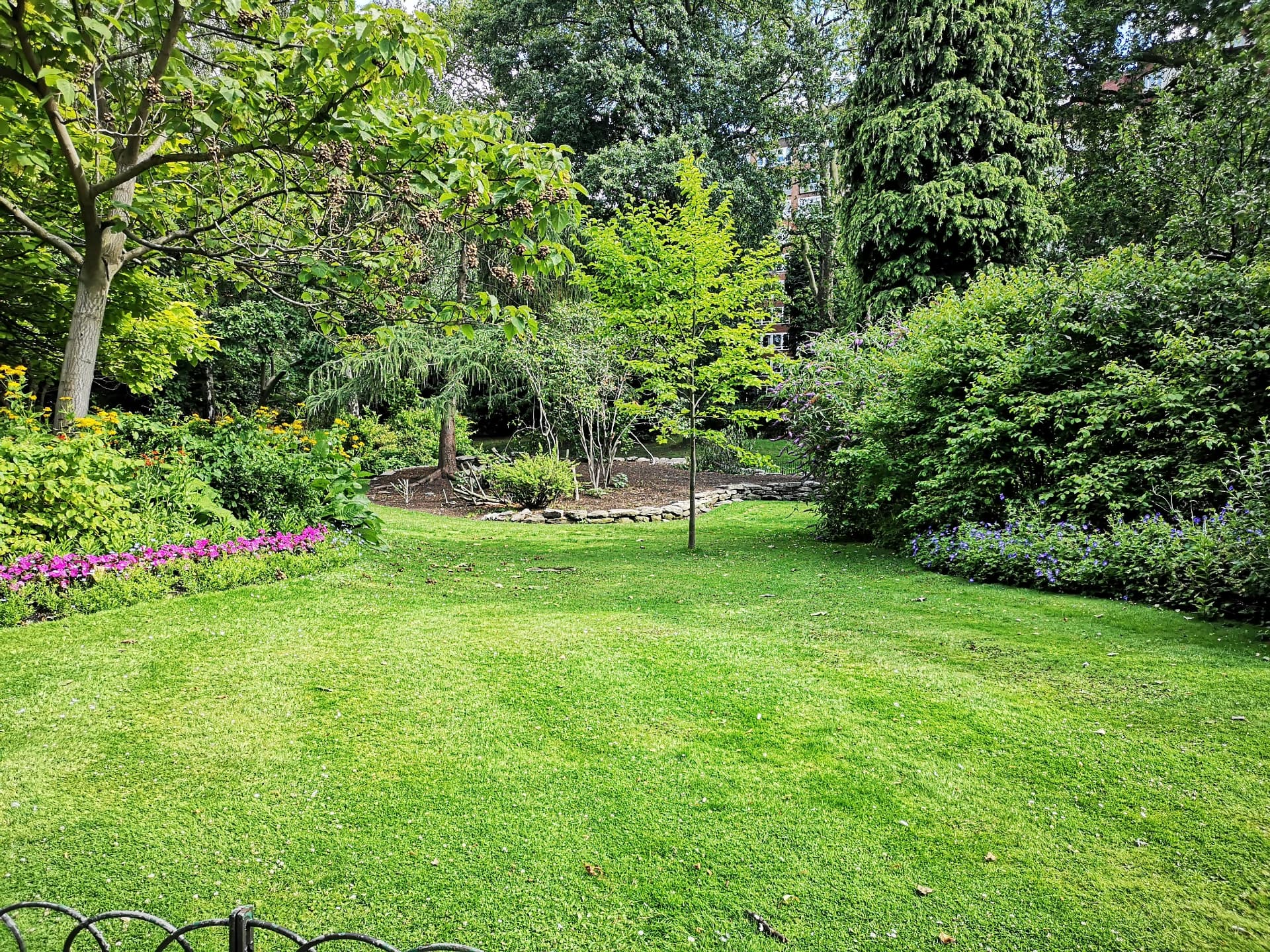 A sunlit lawn framed by mature trees, flowering borders and clipped shrubs.